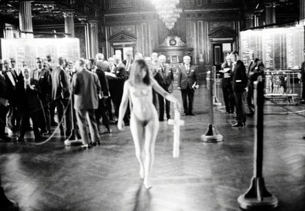 Black-and-white still from VALIE EXPORT’s 1968 performance piece Tap and Touch Cinema. EXPORT walks nude through a large formal interior space while carrying a box-like apparatus strapped to her chest. Groups of formally dressed onlookers stand in the background watching the performance from a distance. Black-and-white still from VALIE EXPORT’s 1968 performance piece Tap and Touch Cinema. EXPORT walks nude through a large formal interior space while carrying a box-like apparatus strapped to her chest. Groups of formally dressed onlookers stand in the background watching the performance from a distance.