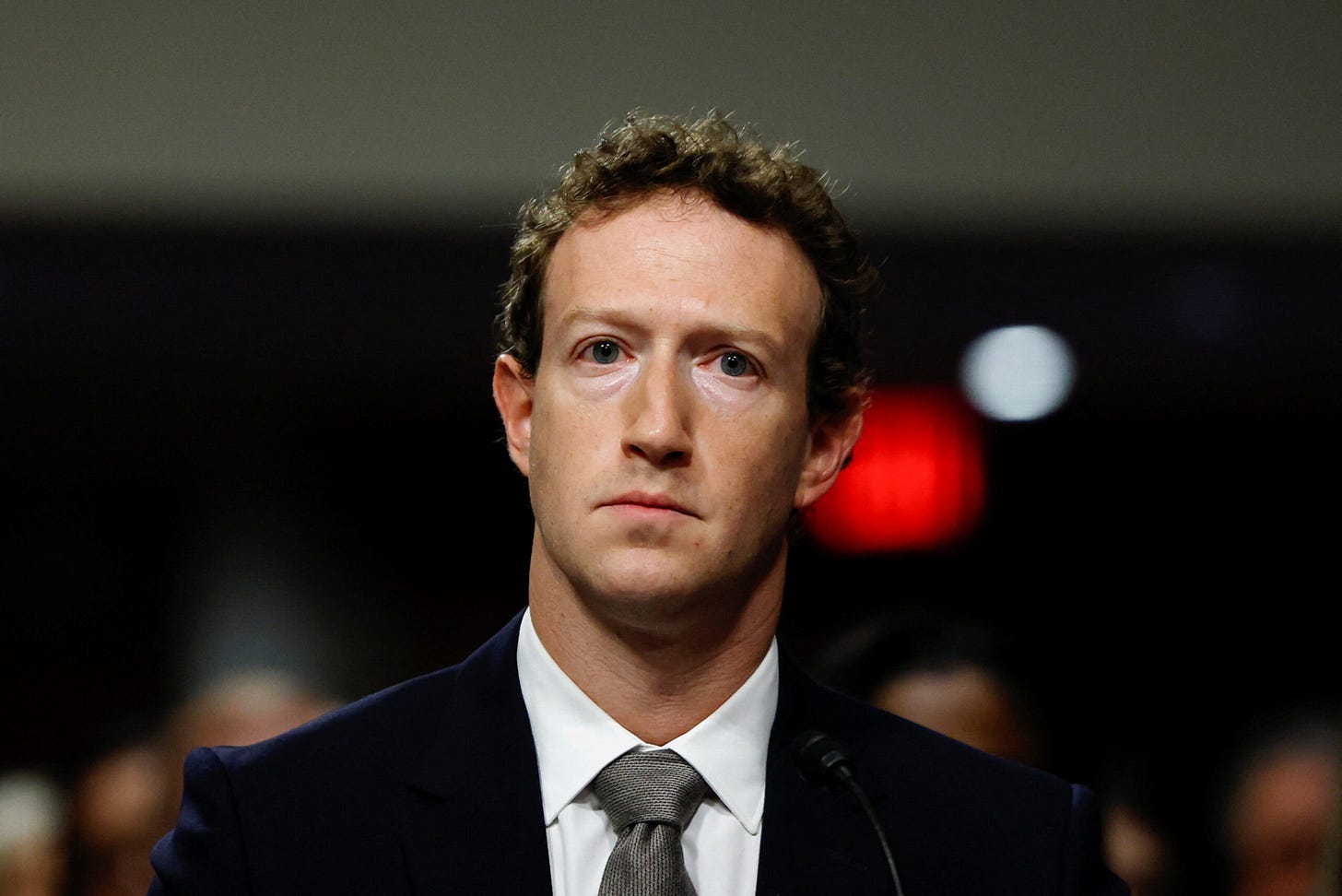 A man with light brown hair and a suit looks wanly at the camera