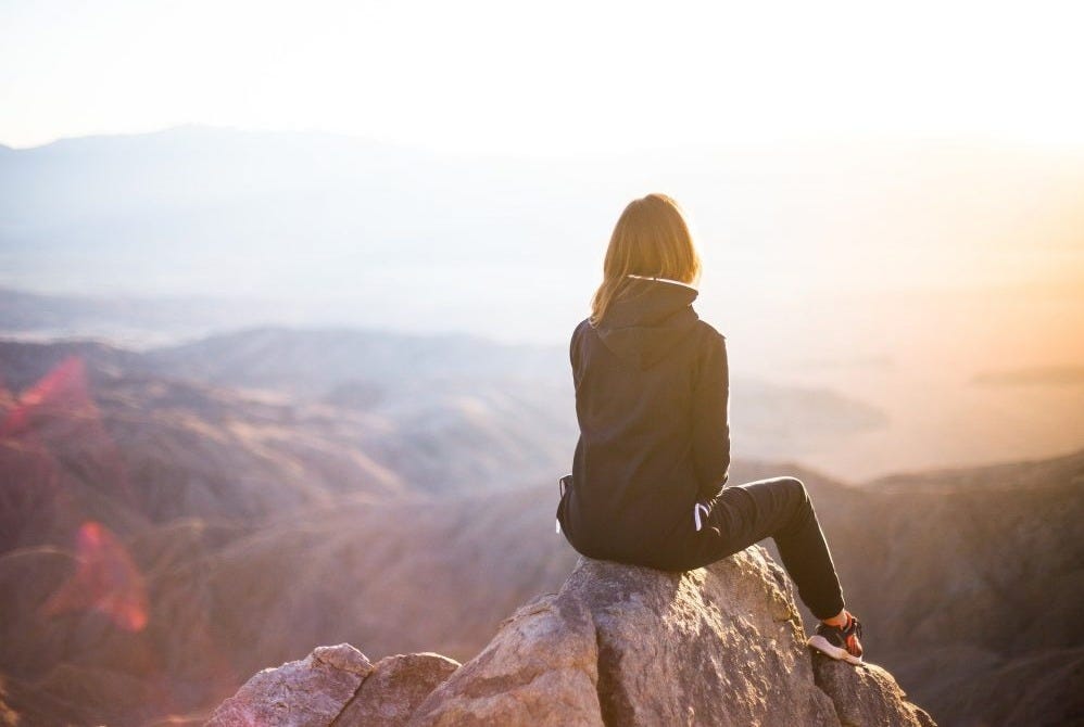 A woman sitting atop a rock at the top of a mountain overlooking other mountain vistas with a bright white obscuring part of the horizon. A woman sitting atop a rock at the top of a mountain overlooking other mountain vistas with a bright white obscuring part of the horizon.