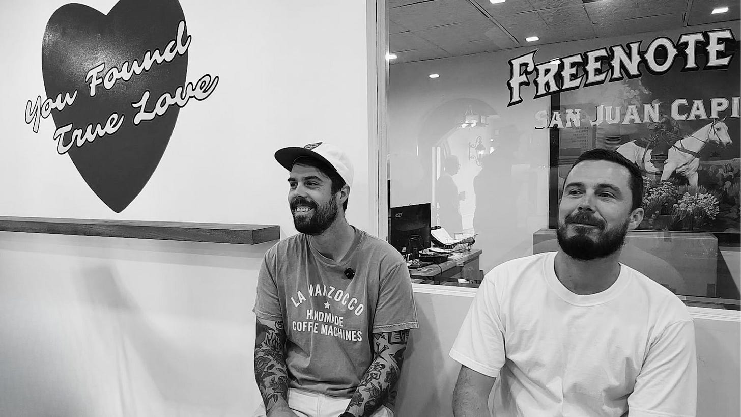 A black and white photo of brothers sitting on a bench with sly grins looking in opposite directions. They're sitting in front of a window inset on a white wall.