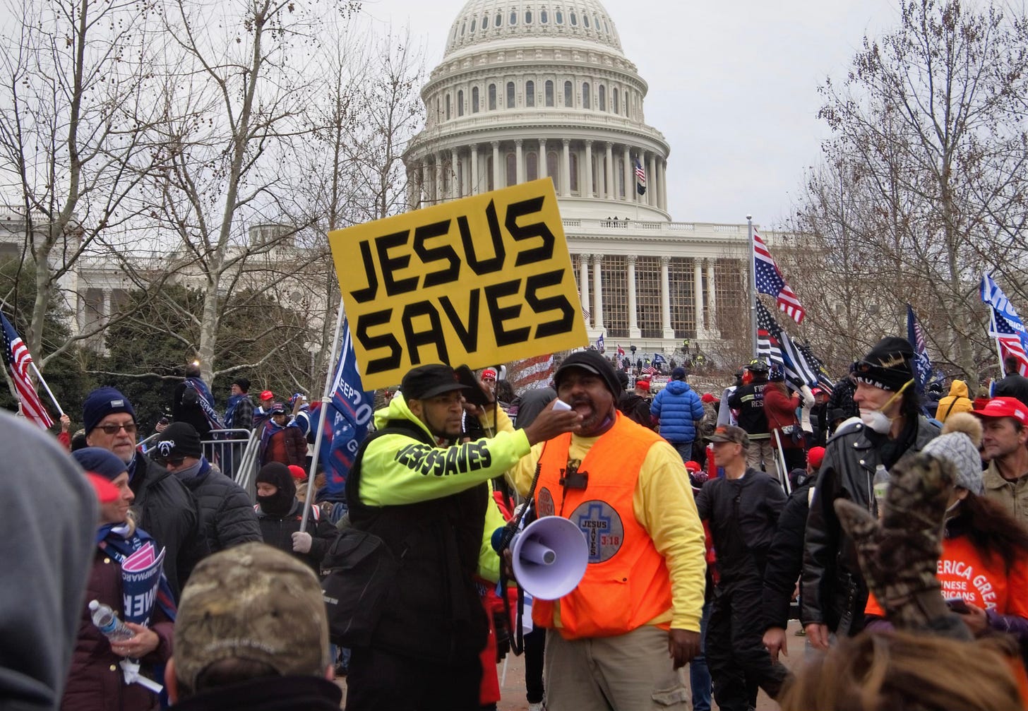 In the midst of the assault on the U.S. Capital on January 6, 2021, two men stand in the midst with bullhorns. One holds a sign that says, "Jesus Saves." The other's shirt says the same, while a large cross is on his companion's vest. The seem to be part of the mob storming the Capital building. In the midst of the assault on the U.S. Capital on January 6, 2021, two men stand in the midst with bullhorns. One holds a sign that says, "Jesus Saves." The other's shirt says the same, while a large cross is on his companion's vest. The seem to be part of the mob storming the Capital building.