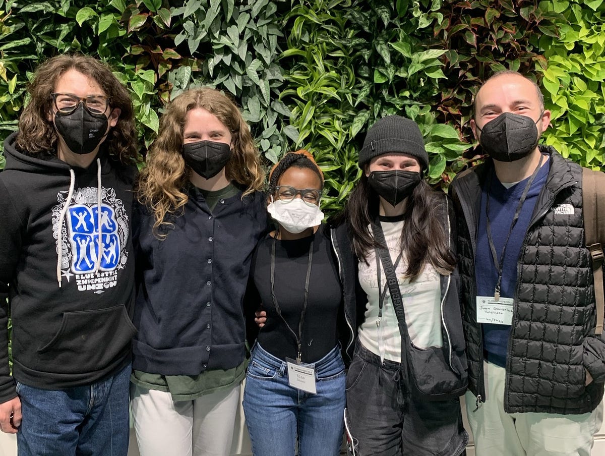 Five people, dressed casually, are standing in front of a wall of plants. The five people are all masked and smiling with their eyes.