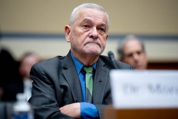 Dr. David Morens with arms crossed at a congressional hearing, wearing a gray jacket, blue shirt, and green tie. 