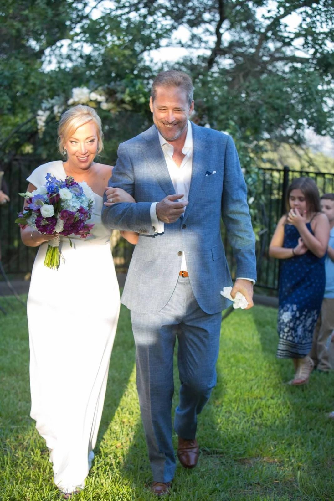Kelly and Nigel walk arm in arm through the garden at their Austin wedding, smiling as guests watch from behind them. Kelly and Nigel walk arm in arm through the garden at their Austin wedding, smiling as guests watch from behind them.