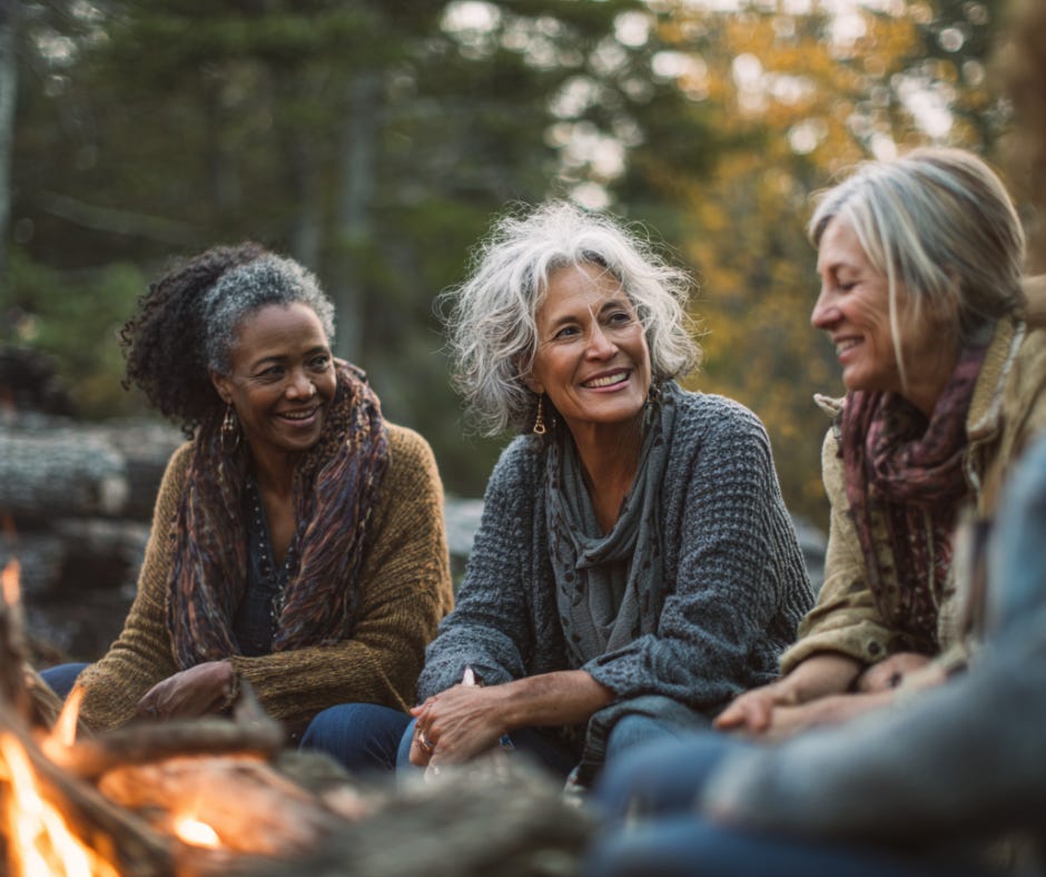 Three older women sit together by a campfire in the woods, smiling and talking, wrapped in cozy autumn sweaters and scarves.