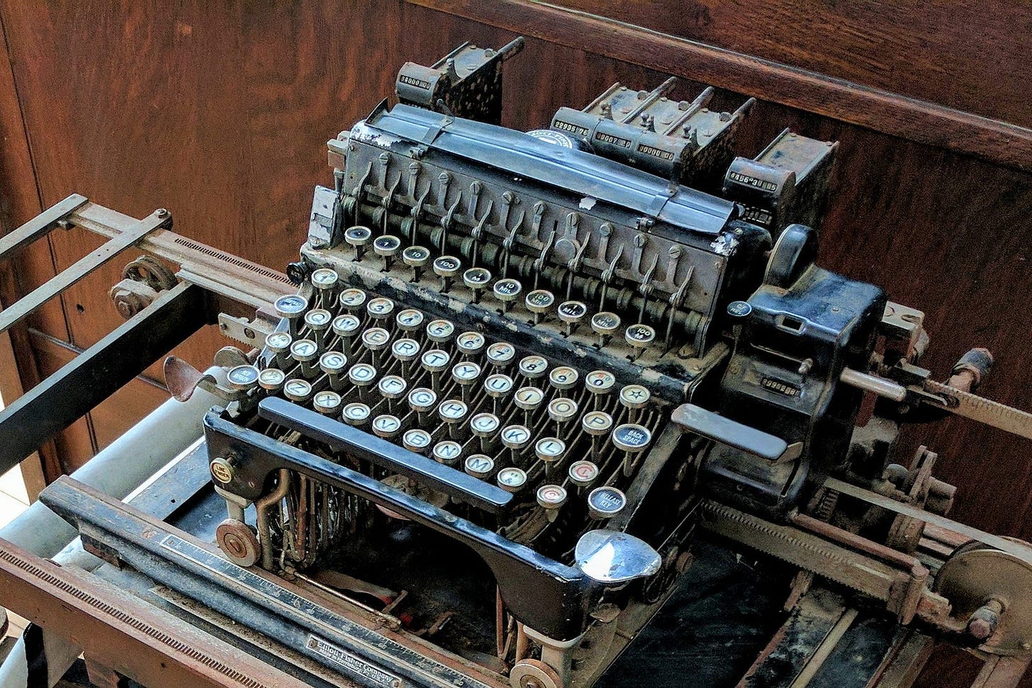A vintage black metal typewriter, possibly an Elliott-Fisher bookkeeping machine from the early 1900s, photographed from above. The typewriter features round glass keys with white letters arranged in a curved QWERTY layout, exposed mechanical typebars, a wide carriage with metal guide rails, and numerical tabulator levers at the top. The machine shows significant age with worn and oxidized metal surfaces. It sits within a wooden desk or cabinet with rich brown wood paneling visible in the background. Photo by the author.