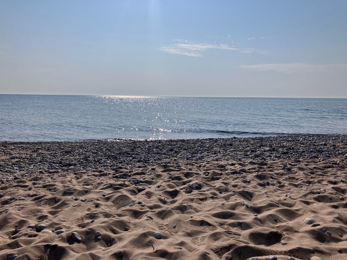 A photo of Lake Michigan. A sandy beach in the foreground, with stones closer to the shoreline. The water is a blue-gray. There are no waves, just ripples. The sky is a light blue, and there is a wispy cloud in a clear, sunny sky.