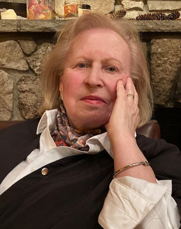 Ms. Hoelterhoff, with blonde hair and wearing a scarf around her neck under a white shirt and dark cardigan, rests her hand against her face.