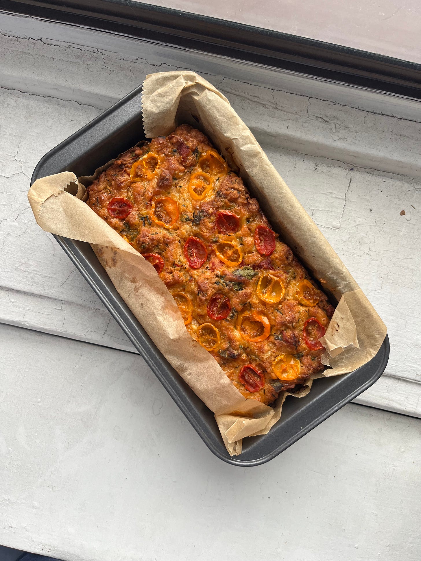 A loaf of zucchini bread sits in a loaf pan on a windowsill. The loaf has halves of red and yellow cherry tomatoes pressed into the top.