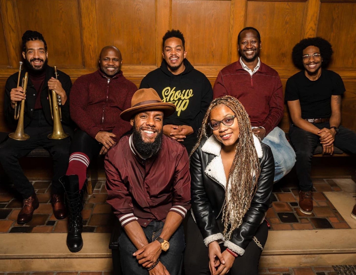 The group of Joe Davis & The Poetic Diaspora sit in a darker room smiling towards the camera The group of Joe Davis & The Poetic Diaspora sit in a darker room smiling towards the camera