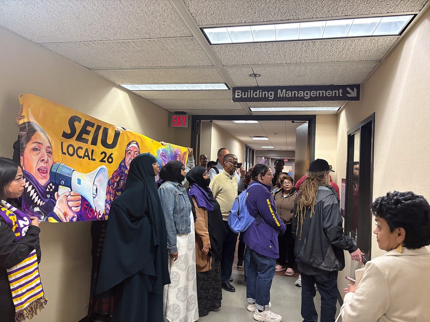 a group of people carrying an SEIU Local 26 banner stand in front of a Building Management office a group of people carrying an SEIU Local 26 banner stand in front of a Building Management office