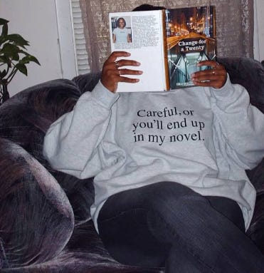 Shamontiel holding her "Change for a Twenty" book in front of her face with a "Careful, or you'll end up in my novel" sweatshirt