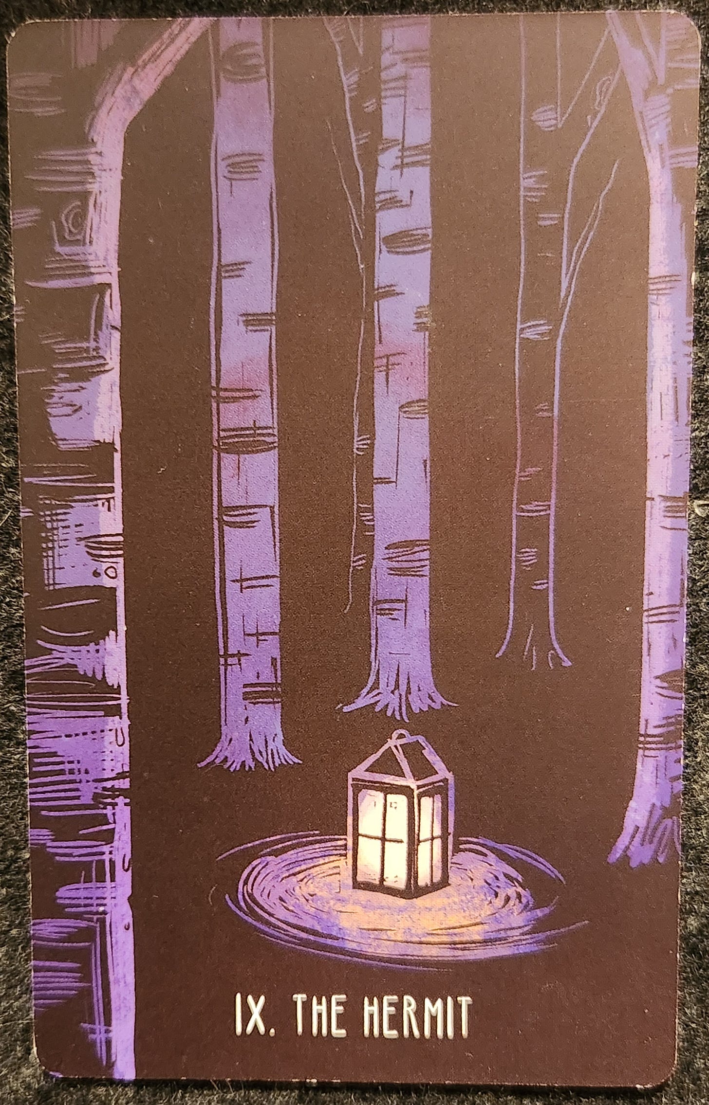 A tarot card labelled IX The Hermit. A lit lantern sits in the middle of a forest clearing and casts a circle of light around itself. It is the only source of light in the dark forest. Around it, tall slender trees reach up to the sky. 