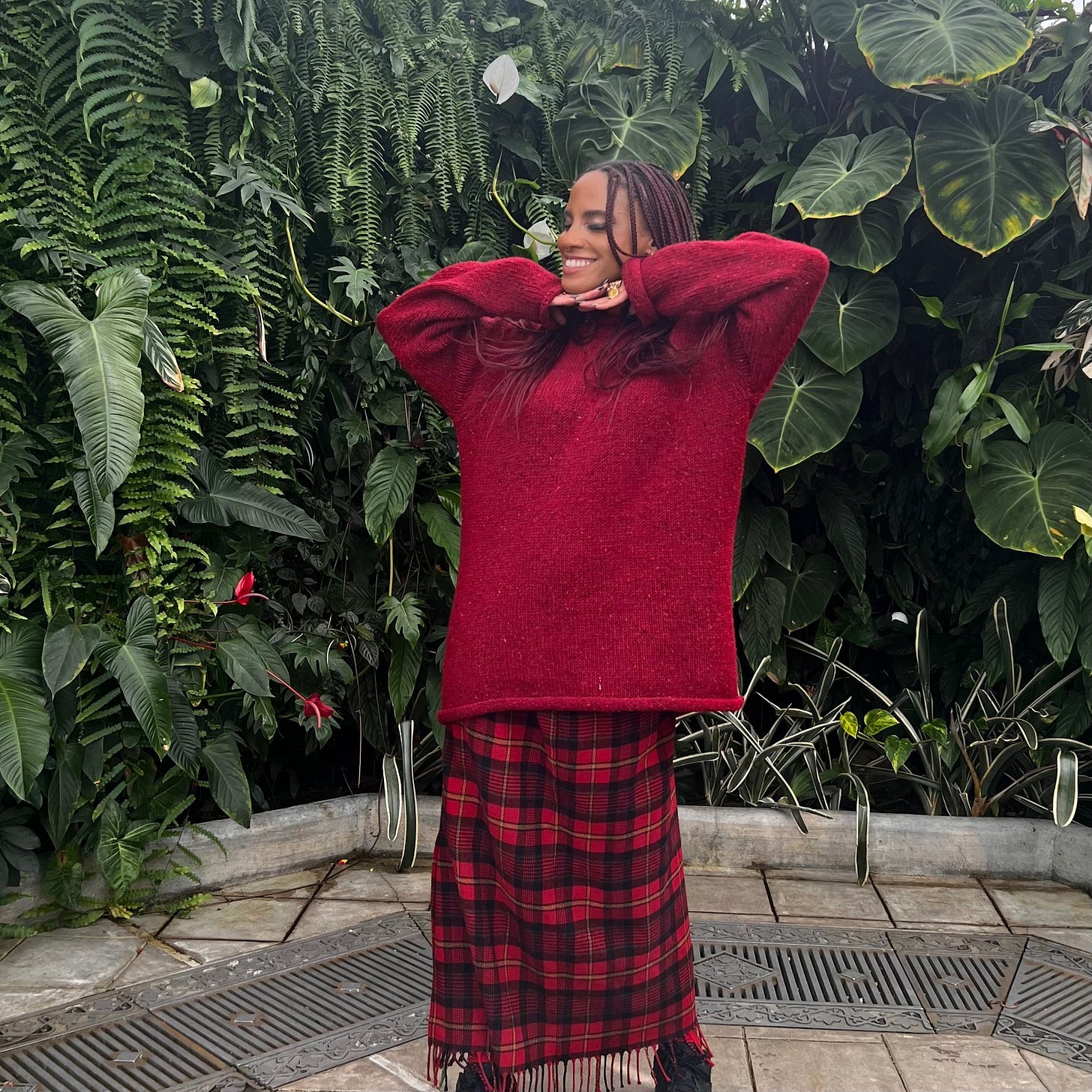 ryan elizabeth peete smiling in a red sweater and plaid skirt standing in front of lush tropical plants inside the Conservatory of Flowers in San Francisco