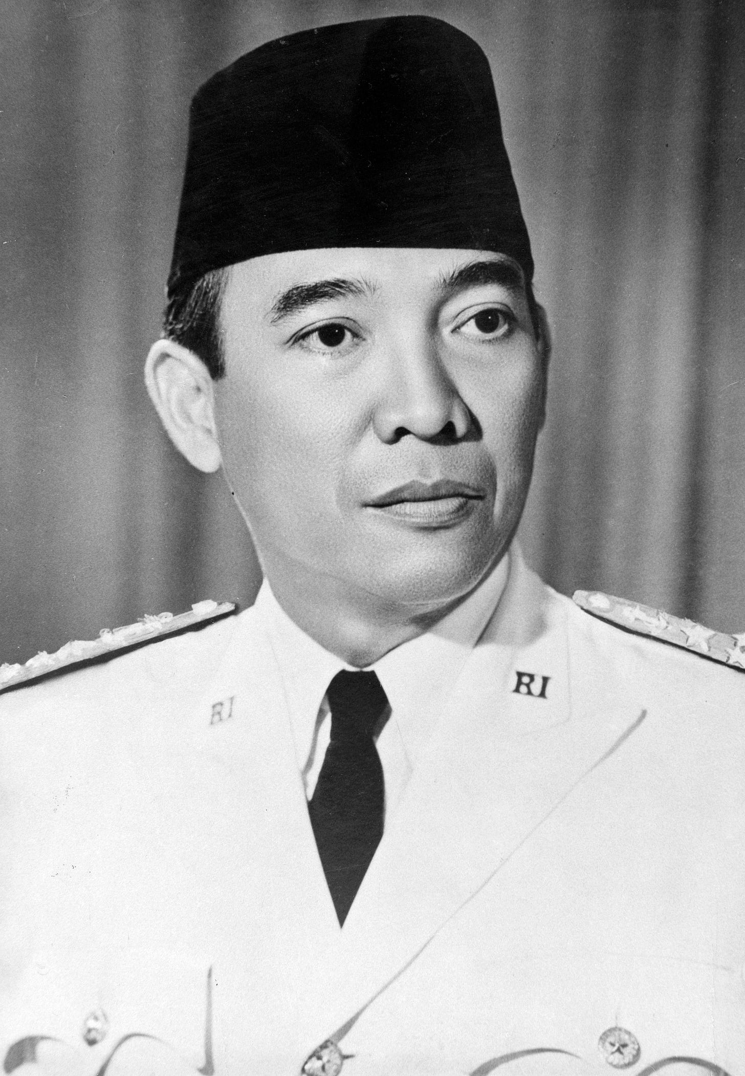 Sukarno is shown from the chest up, his face in three-quarters profile, in front of a curtain backdrop. He wears military dress whites, a black tie, and a black cap known as a songkok or a kopiah (a traditional cap worn by Muslim men in Southeast Asia).