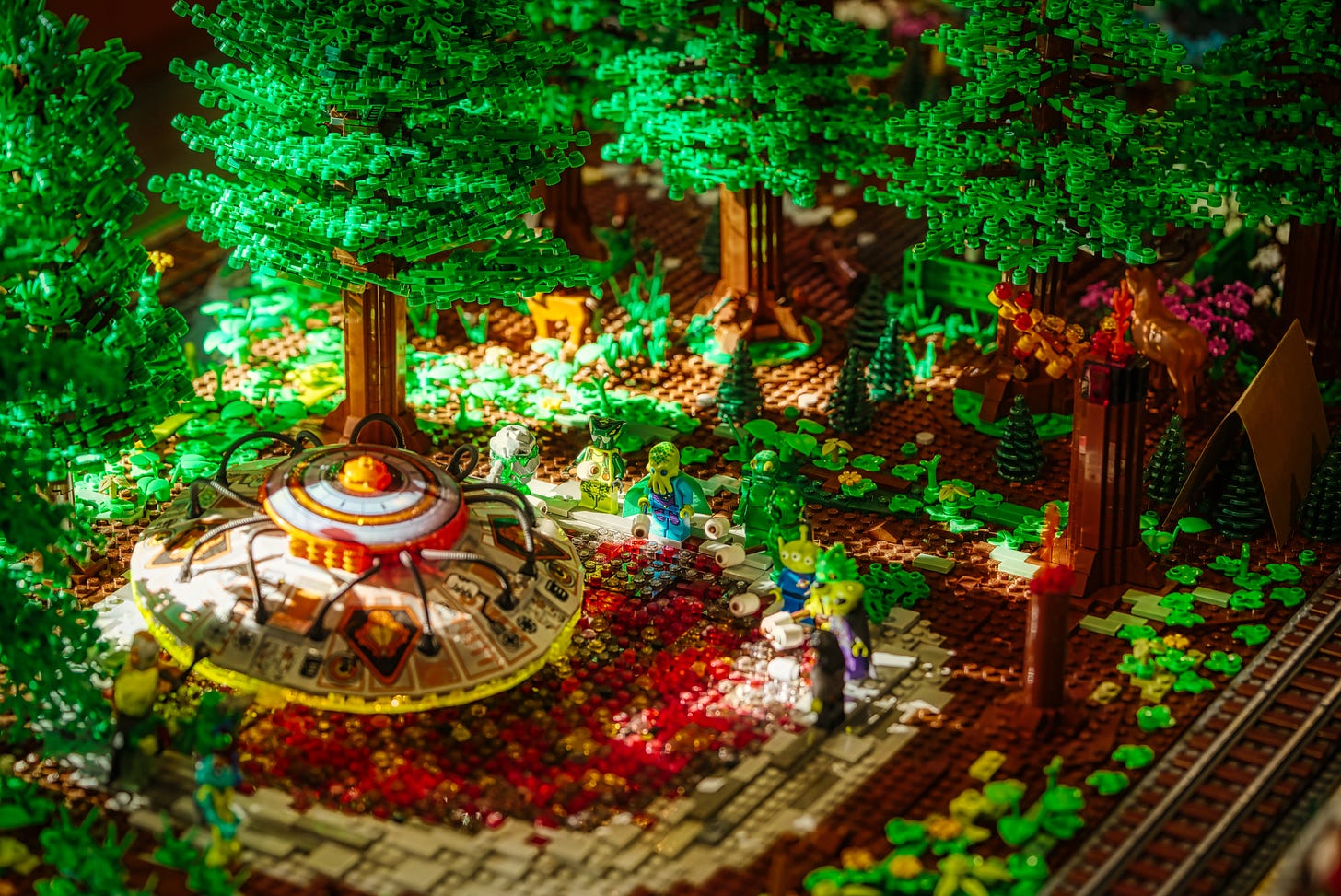 LEGO diorama of a dense green forest with a domed UFO in the foreground, small alien minifigures by a red translucent stream, and a deer and campfire among the trees.