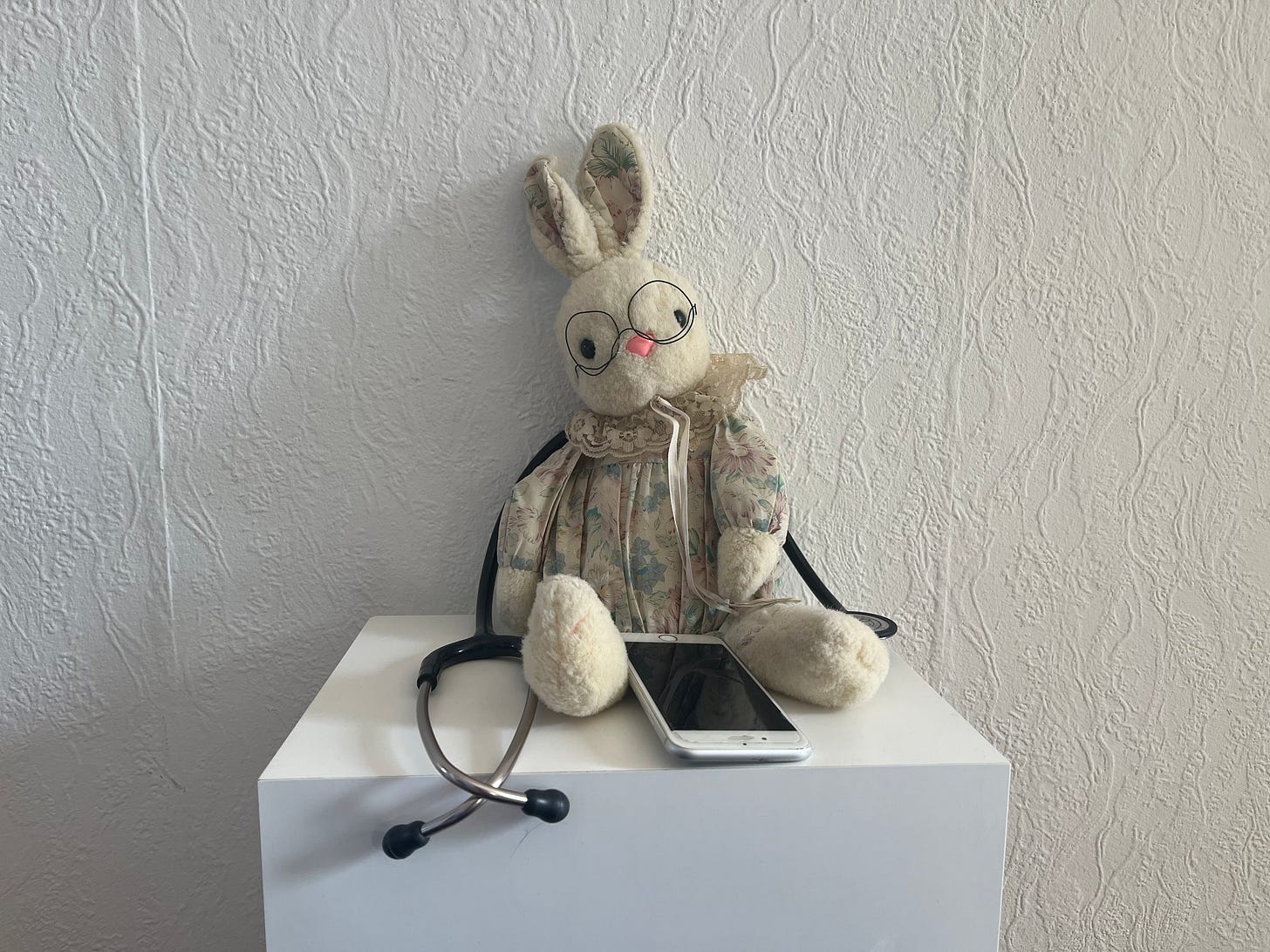 Ein Kuschelhase mit Brille sitzt mit Tablet und Stethoskop vor einer weißen Wand.
