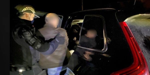 Man with face blurred being arrested and placed in a police car.