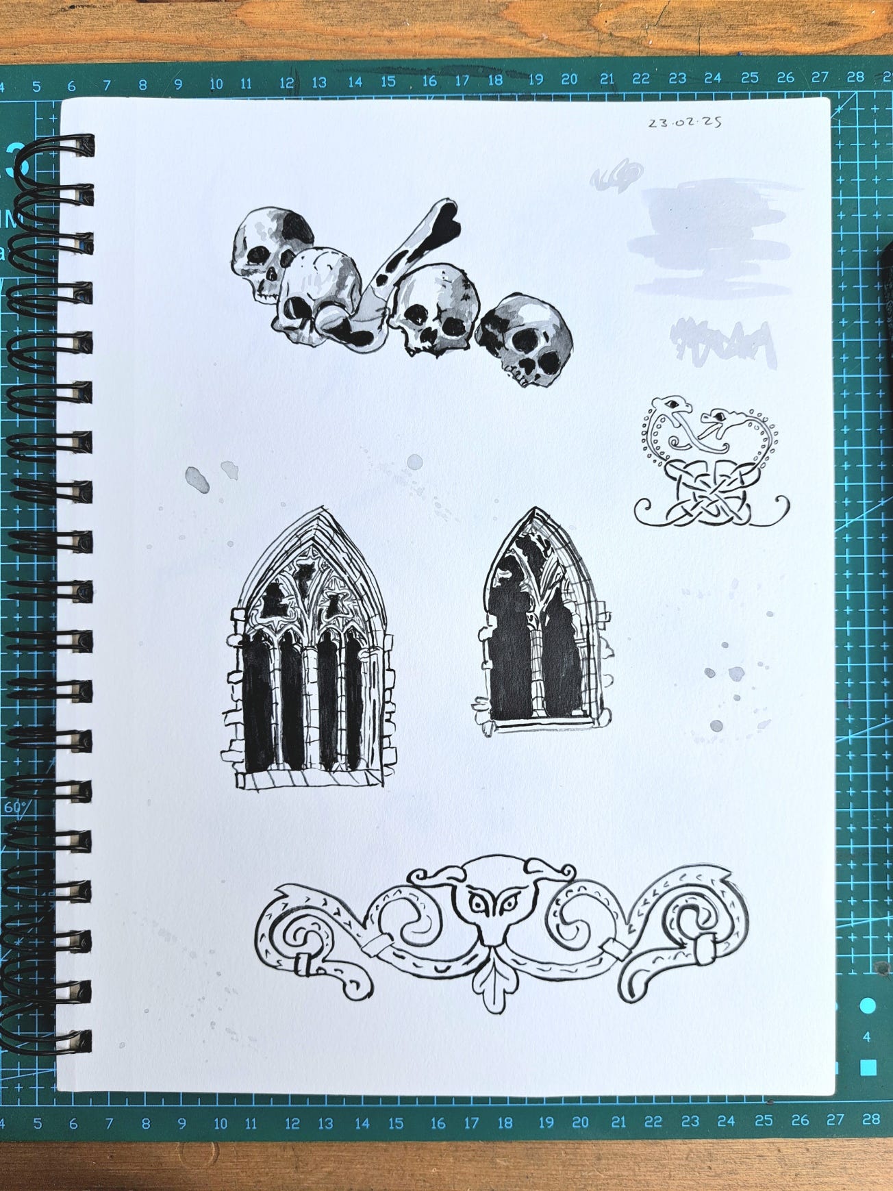 A wirebound sketchbook resting on a green cutting mat. The artworks include skulls, gothic windows and flourishes from medieval manuscripts