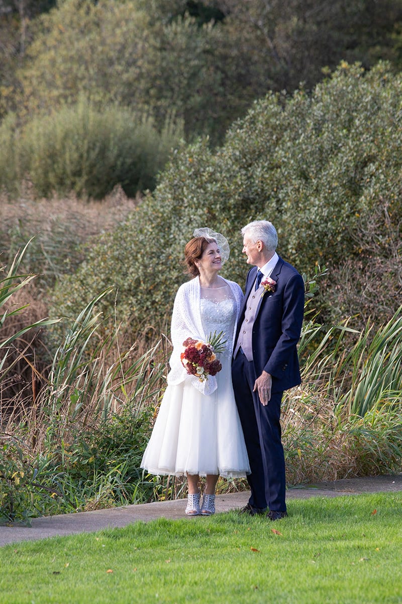 A wedding photograph of Sheena and her husband. He wears a suit with a flower in his lapel; she wears a white dress that stops mid calf, and a veil fascinator. They look into each others eyes, surrounded by greenery.