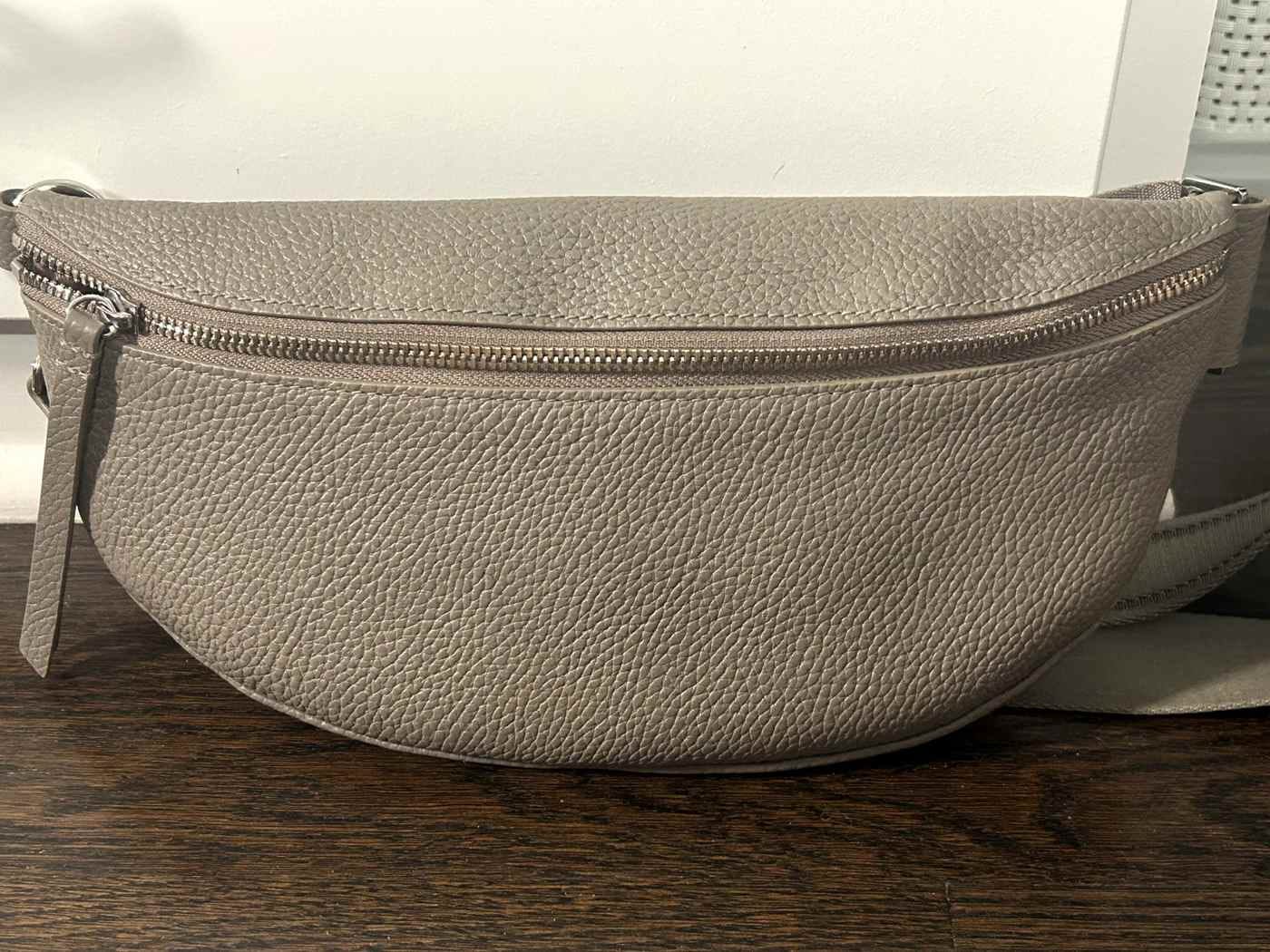 A taupe pebbled-leather Quince sling bag with a silver zipper on a wood table.