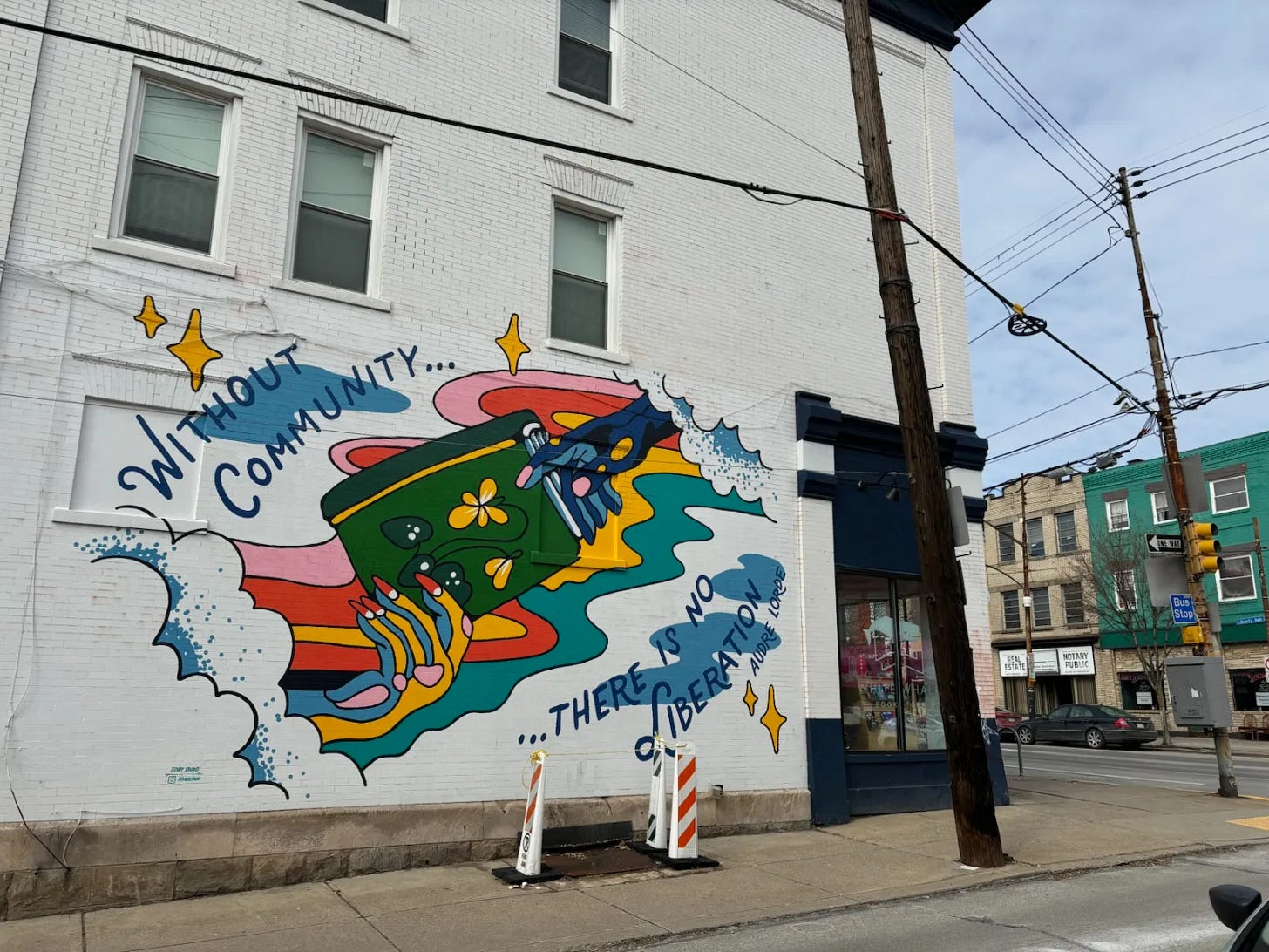 Colorful mural on a white wall of two hands holding a book. Around them are clouds, stars, and the quote "Without community...there is no liberation" by Audre Lorde. The mural is on a building outside. It is daytime.