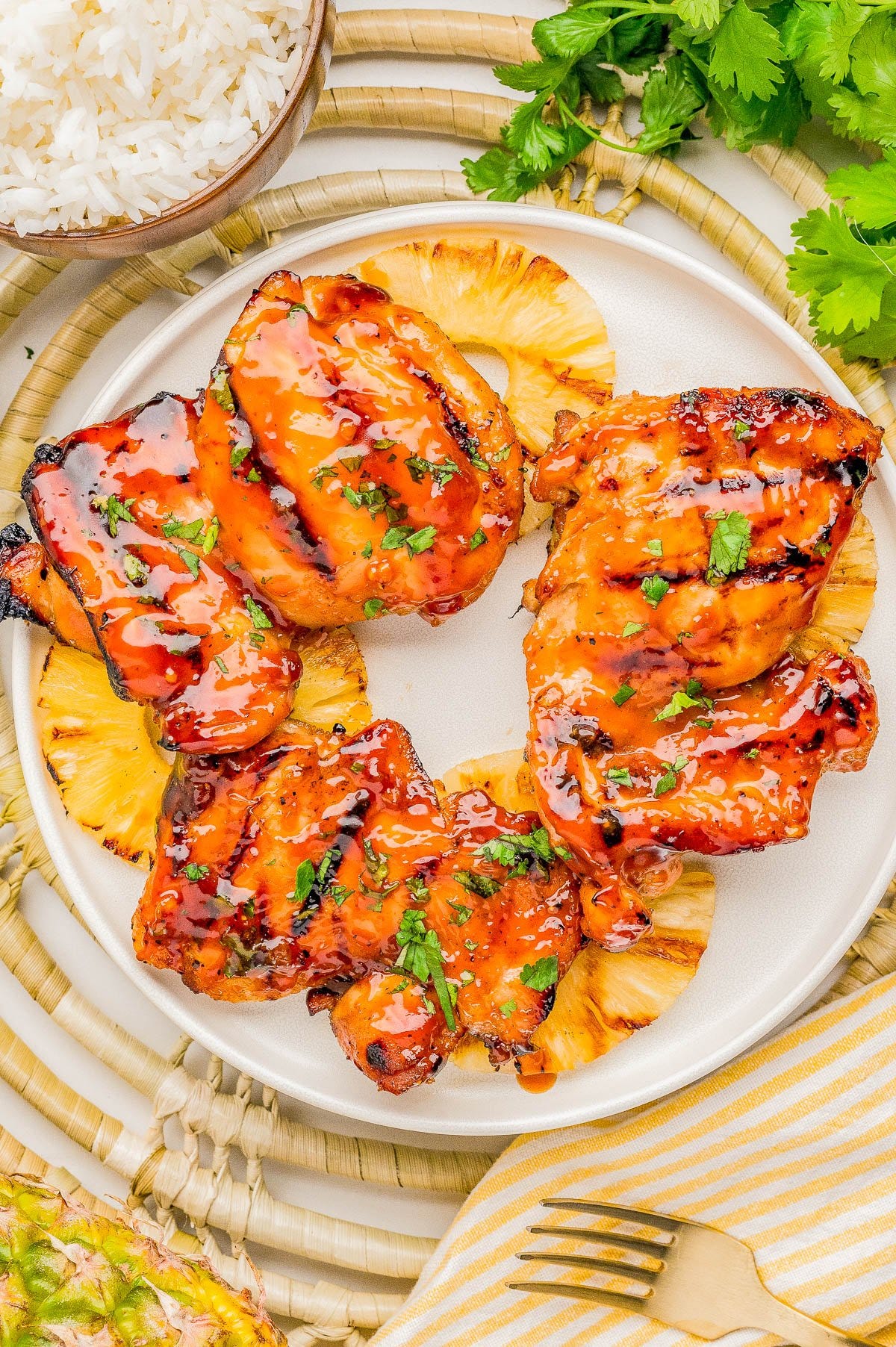 Grilled glazed chicken thighs served on top of pineapple slices, garnished with chopped herbs, on a white plate with rice and fresh cilantro nearby.
