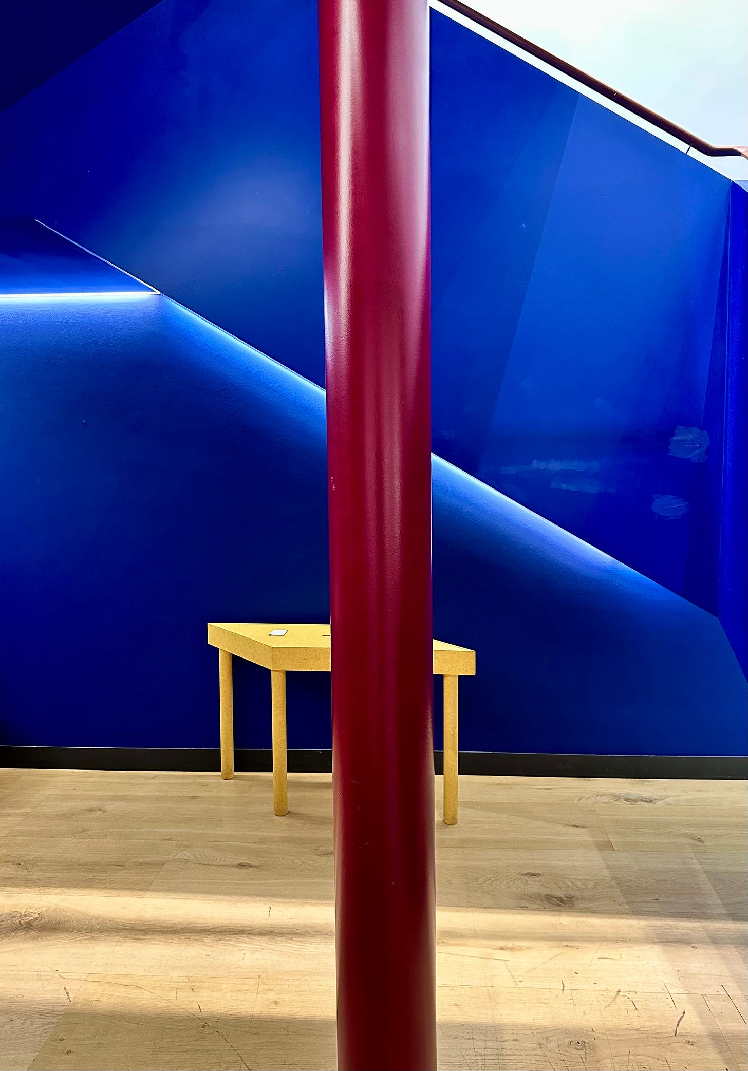 A dark magenta structural support pillar that runs vertically down in a straight line in a room with hardwood floor and intense dark blue wall behind it.