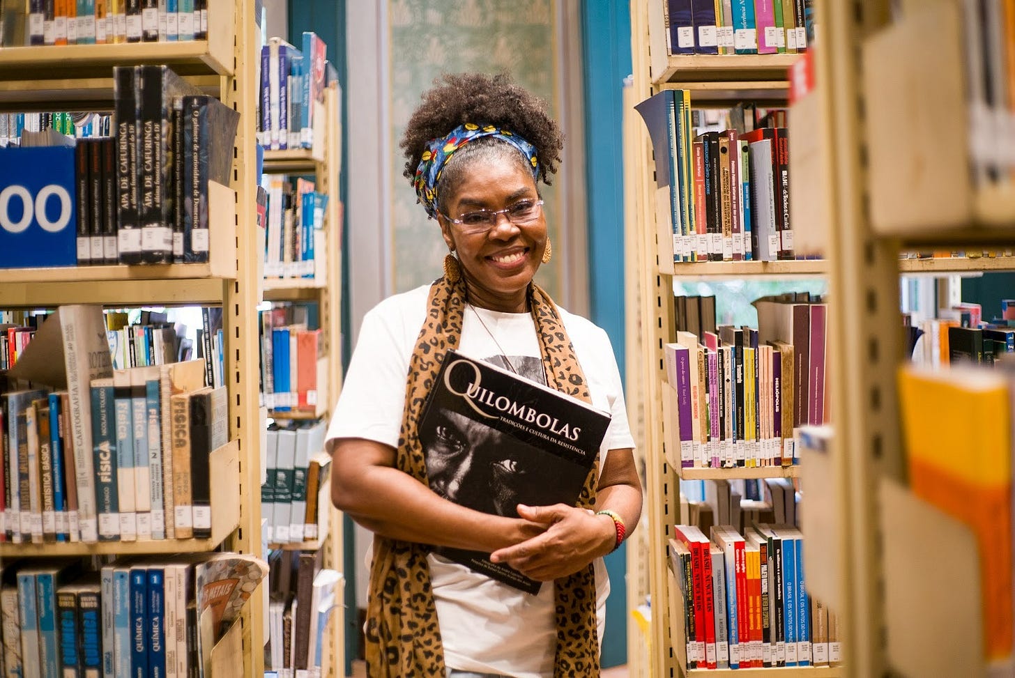 Imagem em ambiente interno, em uma biblioteca. Ao centro, entre duas estantes altas repletas de livros, está Eli Odora, uma mulher adulta, negra, sorridente, voltada para a câmera. Ela usa óculos de grau, brincos médios e um turbante colorido que prende o cabelo crespo, armado no topo da cabeça. Veste uma camiseta branca e um lenço de estampa animal print em tons de marrom e preto, colocado ao redor do pescoço. Nos braços, segura um livro grande, de capa preta, com o título “Quilombolas”, em letras brancas. A capa traz a imagem de um rosto em preto e branco. As estantes ao redor estão organizadas, com livros de diferentes tamanhos e cores, muitos com etiquetas na lombada, indicando catalogação. O corredor entre as prateleiras é estreito e bem iluminado, transmitindo um ambiente acadêmico e silencioso. A expressão da mulher transmite acolhimento, orgulho e satisfação por estar naquele espaço de conhecimento.