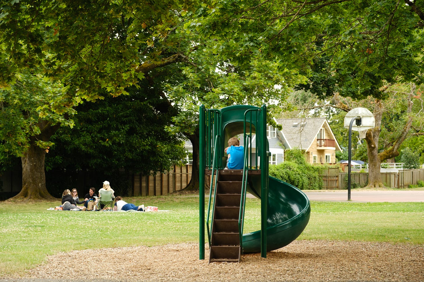 A child wearing a turquise t-shirt sits atop a slide in a city park; nearby, five women sit on the ground and in chairs having a picnic. The backdrop is leafy green maples. 