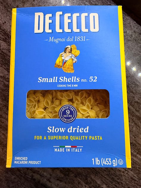 Image 1: A white bowl filled with small shell pasta in a tomato-based broth with vegetables, served on a wooden table. A torn piece of rustic brown bread rests on the rim of the bowl. Image 2: A blue-and-yellow box of De Cecco Small Shells No. 52 pasta, showing the pasta through a clear window on the front. The packaging highlights that it is slow‑dried for superior quality and made in Italy. Image 3: A large pot filled with a simmering mixture of diced vegetables—such as onions, celery, and red peppers—in a seasoned broth, likely the base for a soup or pasta dish.