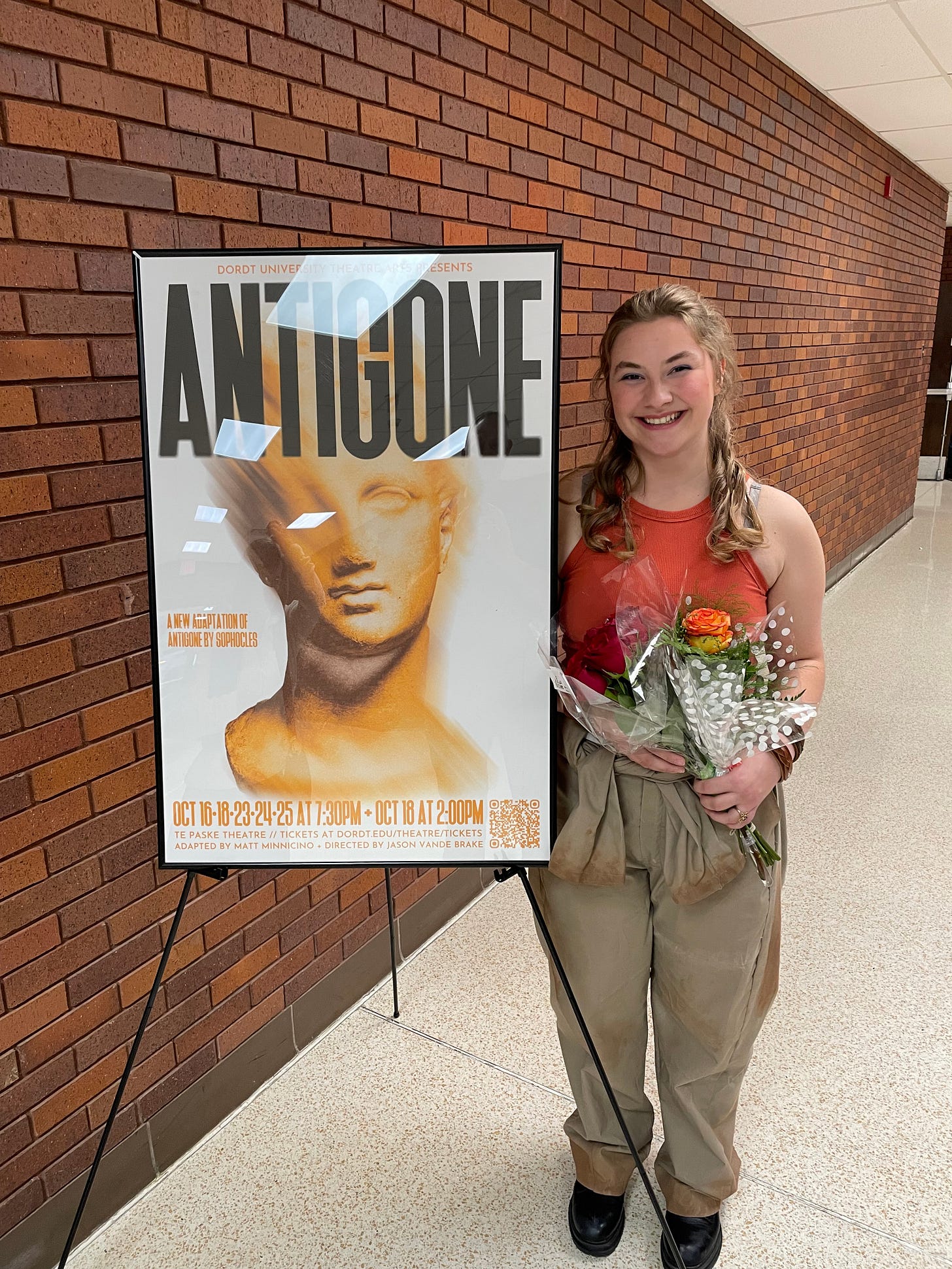 A college thespian next to the poster for the show Antigone