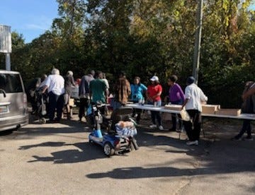 An outdoor scene in a parking lot where a group of people line up at long folding tables, suggesting a community distribution or outreach event. A blue mobility scooter sits in the foreground, a gray minivan is parked to the left, and volunteers and attendees converse along the tables. Trees with autumn foliage and a tall light pole form the background under clear, sunny skies. No recognizable brands or landmarks are visible.