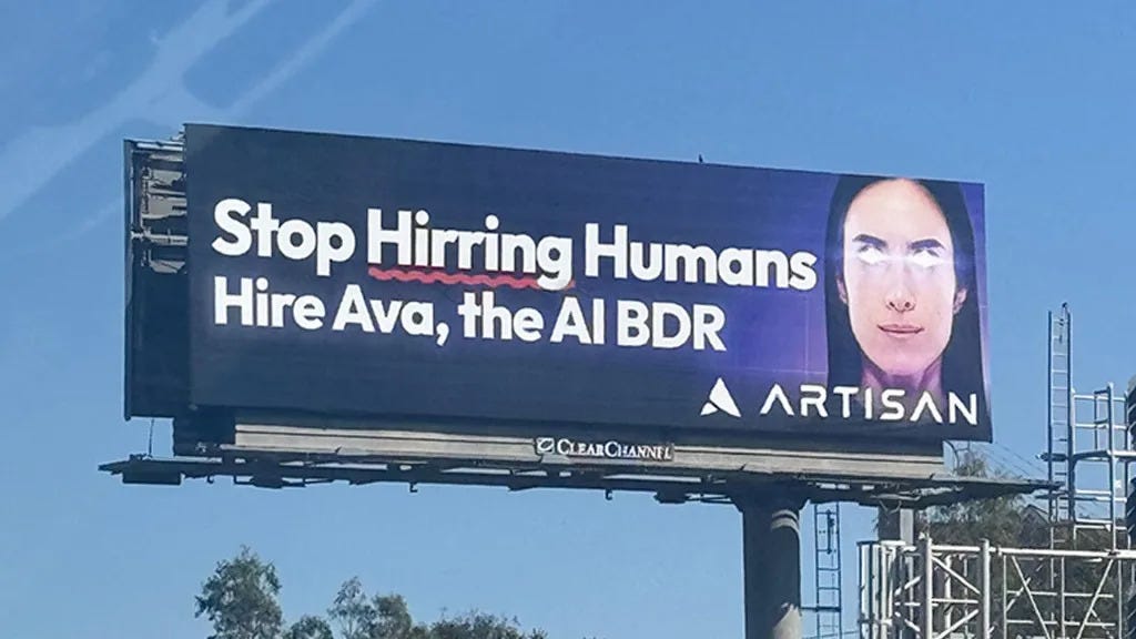 A large roadside billboard reading “Stop Hirring Humans. Hire Ava, the AI BDR” with the word “Hirring” intentionally misspelled and underlined in red. The ad features a stylized image of a woman’s face with glowing eyes on a dark blue background. The billboard promotes Artisan, a company advertising its AI employee product.
