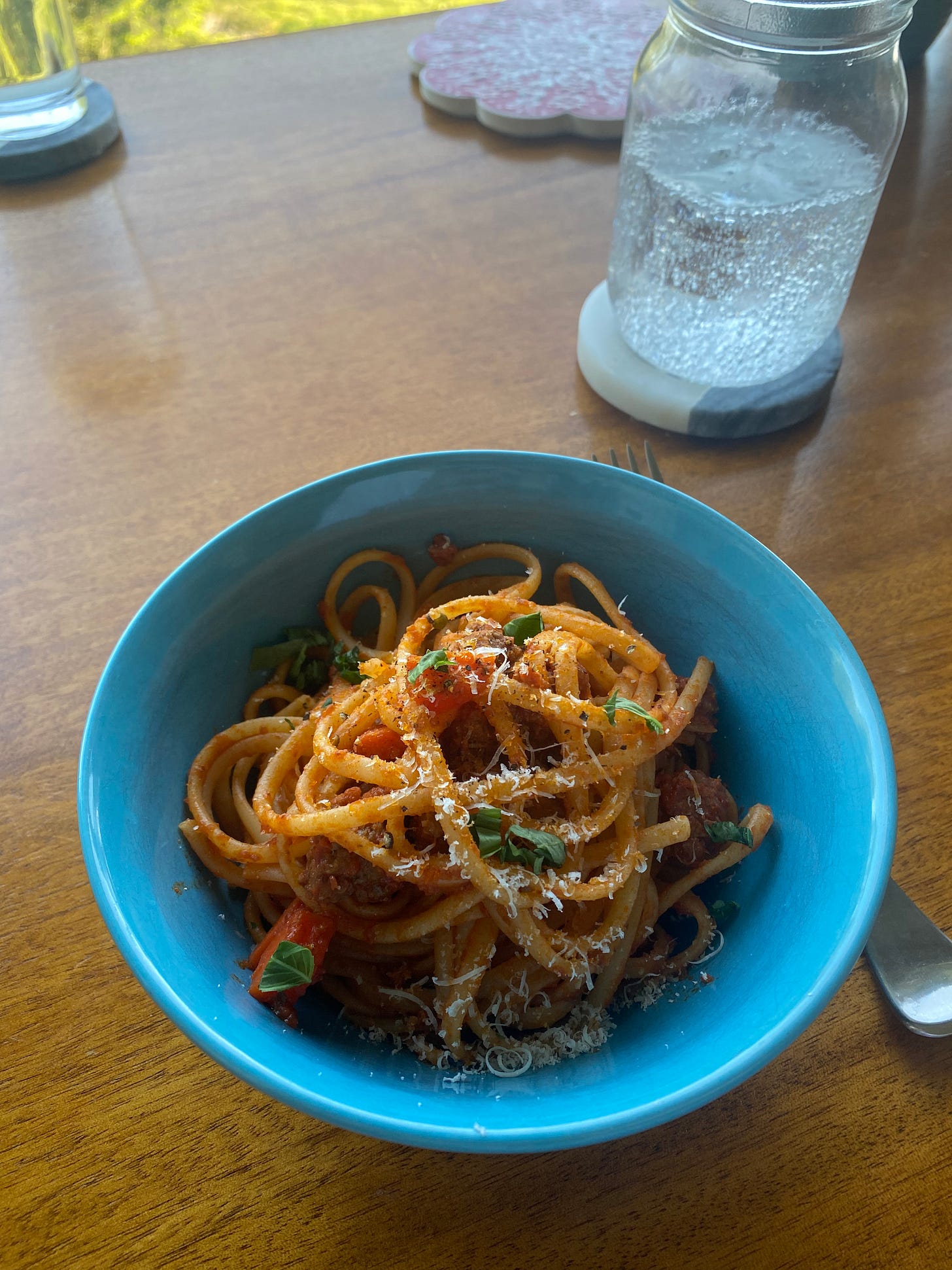A blue bowl of linguine in bolognese sauce, topped with parmesan and fresh basil leaves. A glass of sparkling water is on a coaster in the background.
