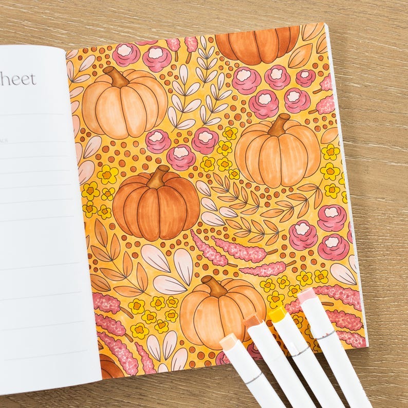 May include: An open notebook with a yellow and orange page featuring a repeating pattern of pumpkins, flowers, and leaves. The page is titled "sheet". Four white markers are lying on a wooden surface next to the notebook.