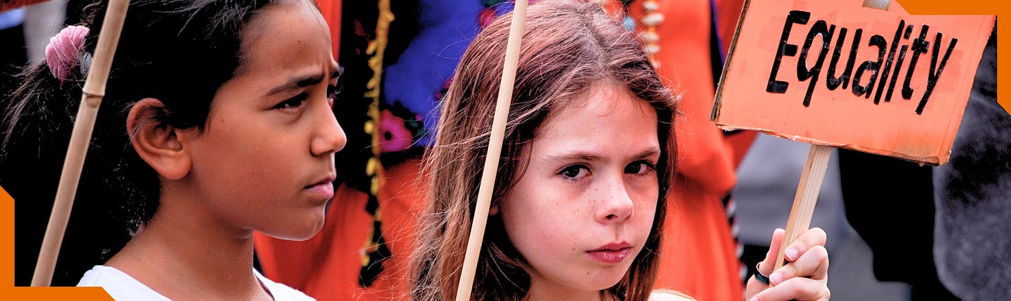 Two children stand at a Women's Day protest rally. The child on the left is looking toward the right of the frame. The child on the right is holding an orange 'Equality' sign and looking straight at the viewer with a serious expression. A crowd wearing vibrant colours is out of focus in the background. 