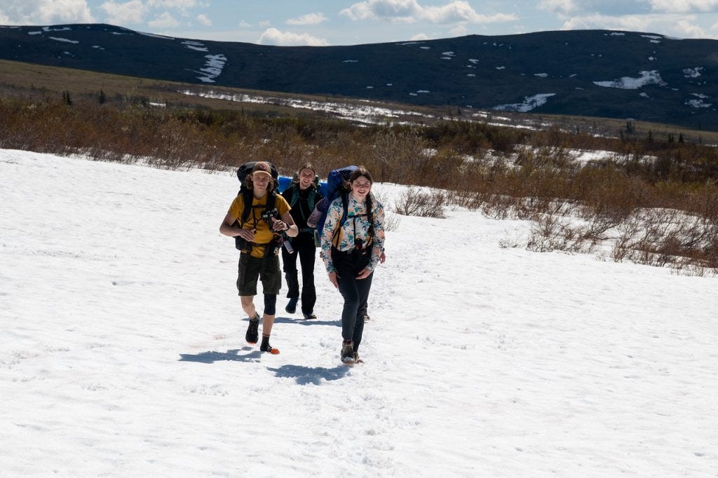 Three hikers with large backpacks walk toward the camera across a broad, sun-bright snowfield, low brush and a long snow-streaked ridge behind them under a blue sky.