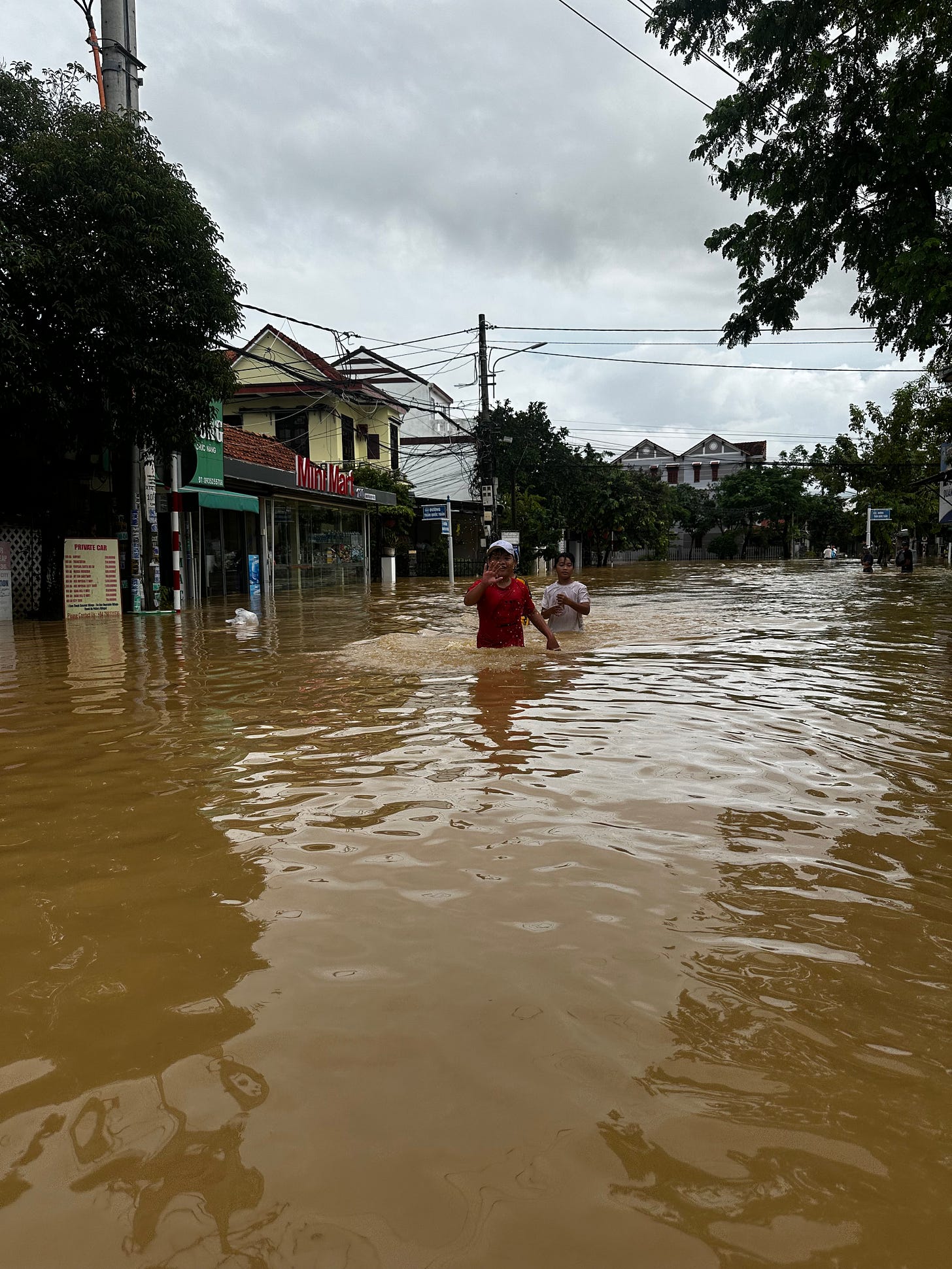 Two kids wade through waist-deep floodwater on a street in Hoi An, Vietnam. The surrounding shops and homes are partially submerged, and the sky is overcast. The kids appear calm, almost jovial, and are looking toward the camera as they move through the water.