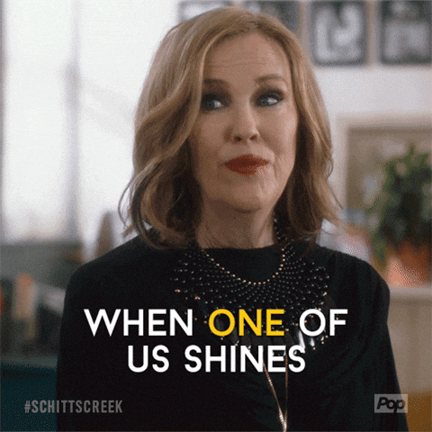 Woman in black shirt with necklace smiles in an interior setting. Text reads "WHEN ONE OF US SHINES." Hashtags and Pop logo visible.