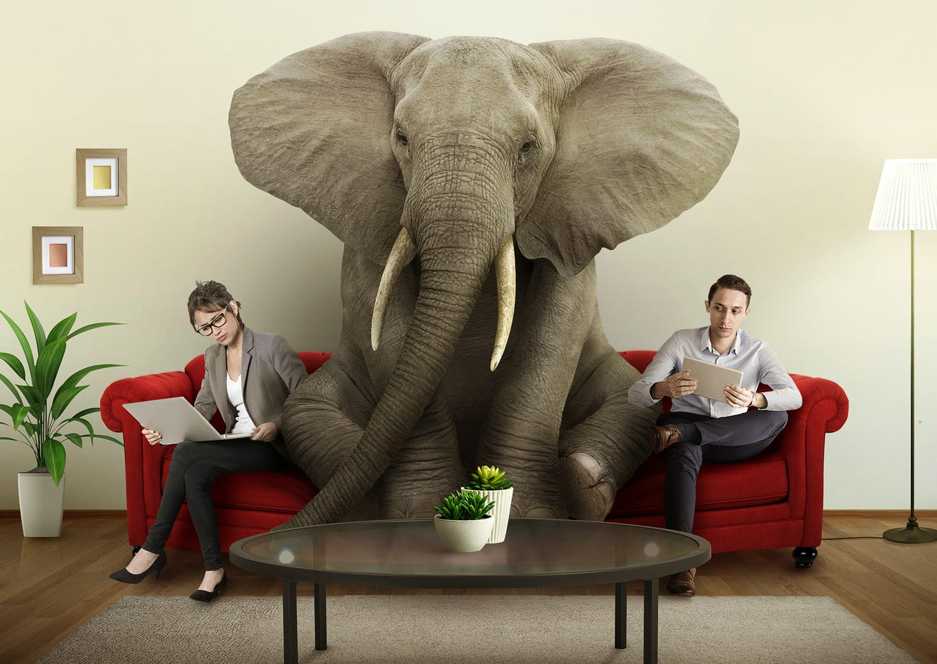 Elephant sits between a woman and man on a red couch busily doing their work, unaware of his presence. Elephant sits between a woman and man on a red couch busily doing their work, unaware of his presence.