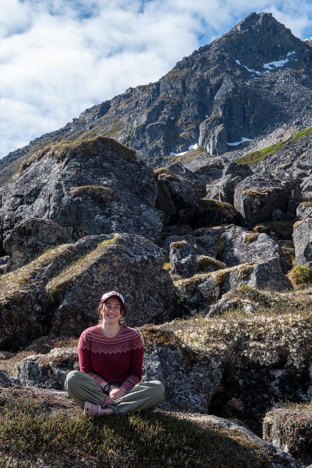 A woman sits cross-legged on a moss- and lichen-covered boulder, smiling at the camera, wearing a dark red patterned sweater, olive pants, and a red and white cap. Behind her, large lichen-spotted boulders pile up a slope that rises to a sharp grey peak streaked with small snow patches, under a blue sky with white cloud. A woman sits cross-legged on a moss- and lichen-covered boulder, smiling at the camera, wearing a dark red patterned sweater, olive pants, and a red and white cap. Behind her, large lichen-spotted boulders pile up a slope that rises to a sharp grey peak streaked with small snow patches, under a blue sky with white cloud.