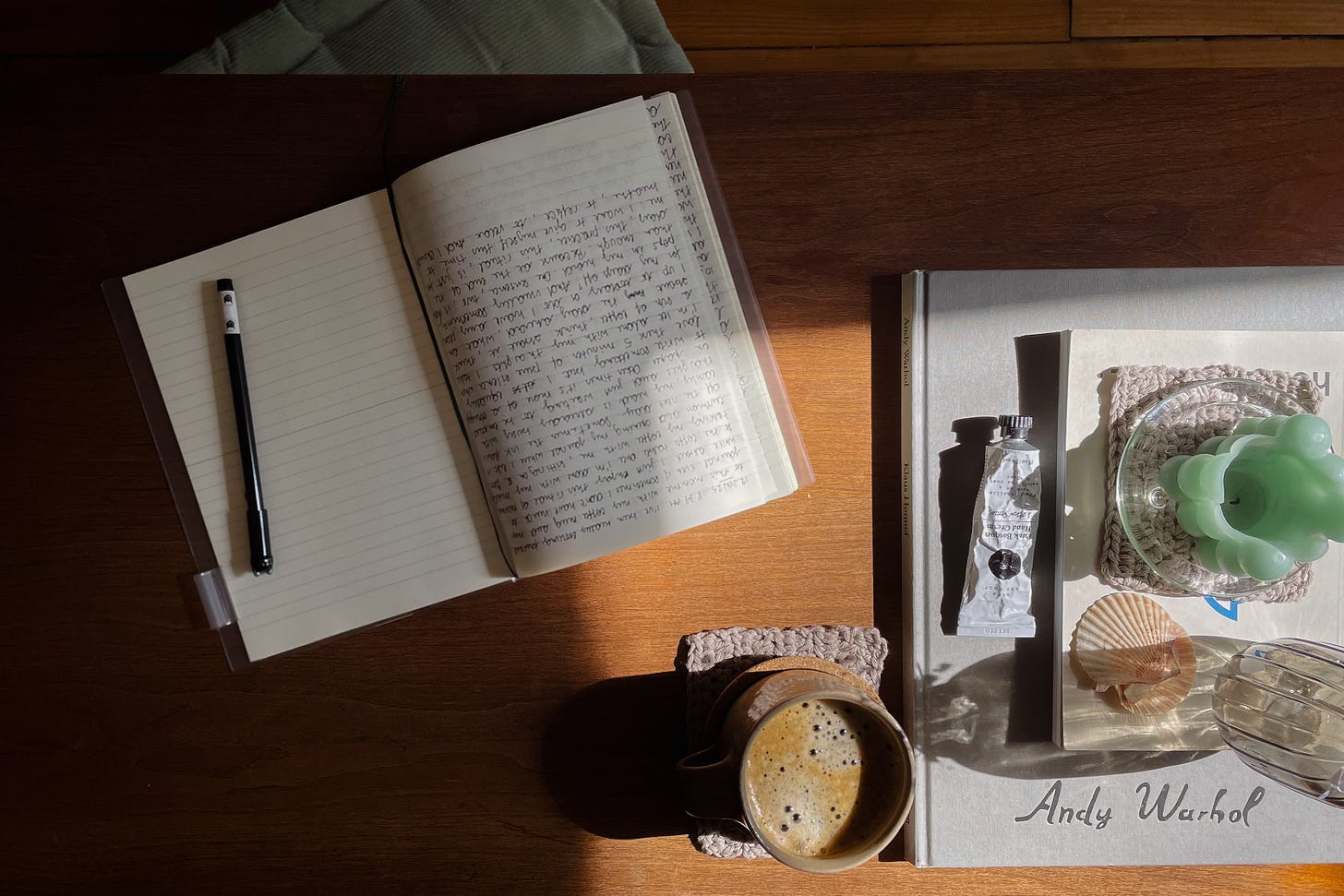 Sunlit wooden coffee table with open journal, pen, coffee cup, and floor cushion below.