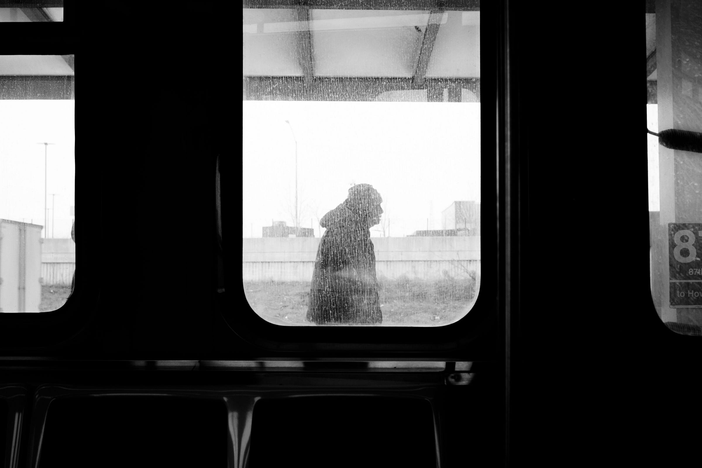 Black-and-white photo of man walking on "L" platform. Photo perspective is looking from the inside of the train through a grimy window.