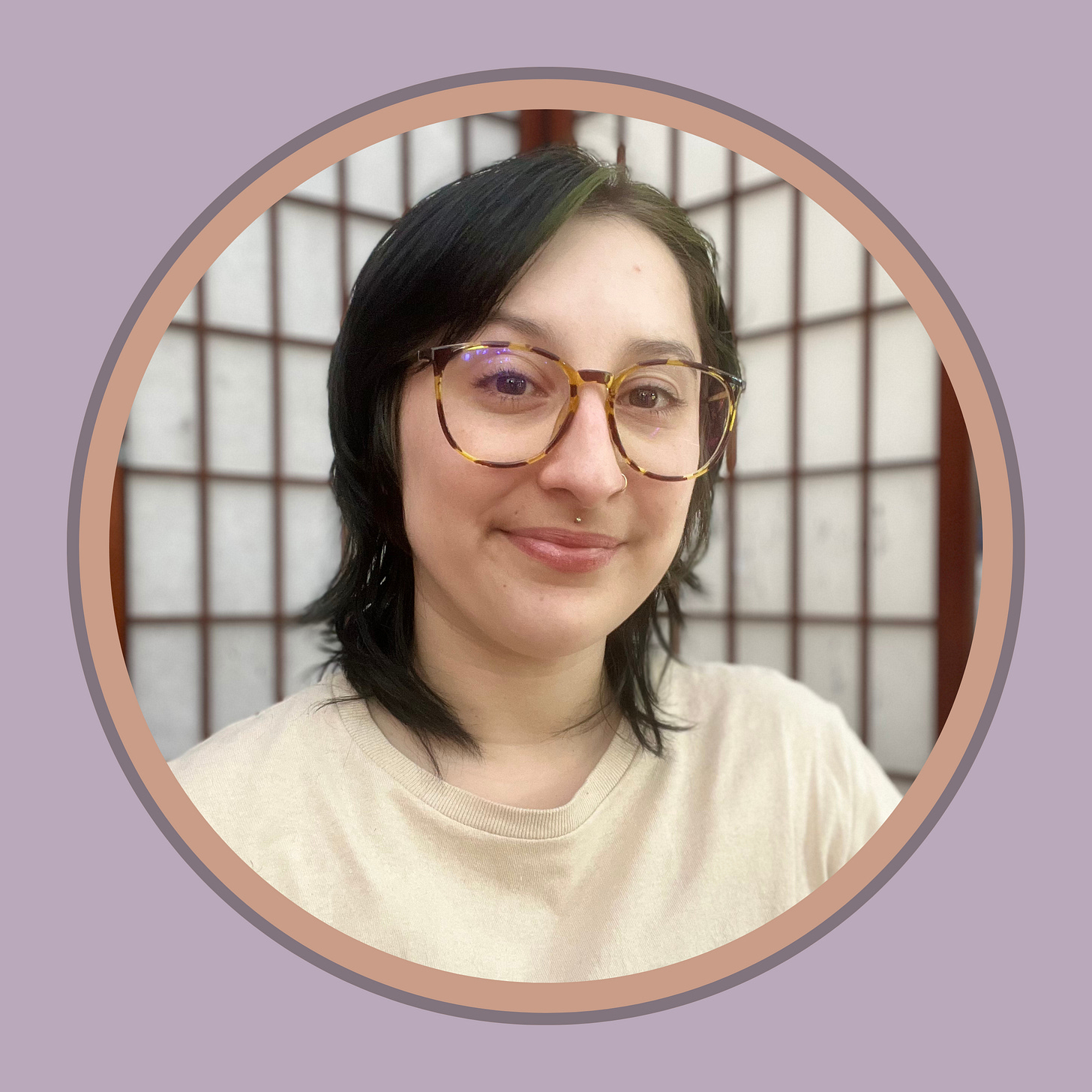 A non binary, femme presenting individual, wearing tortoise shell glasses, has dark, medium length hair, and a cream colored shirt on. They are smiling gently, gazing into the camera.