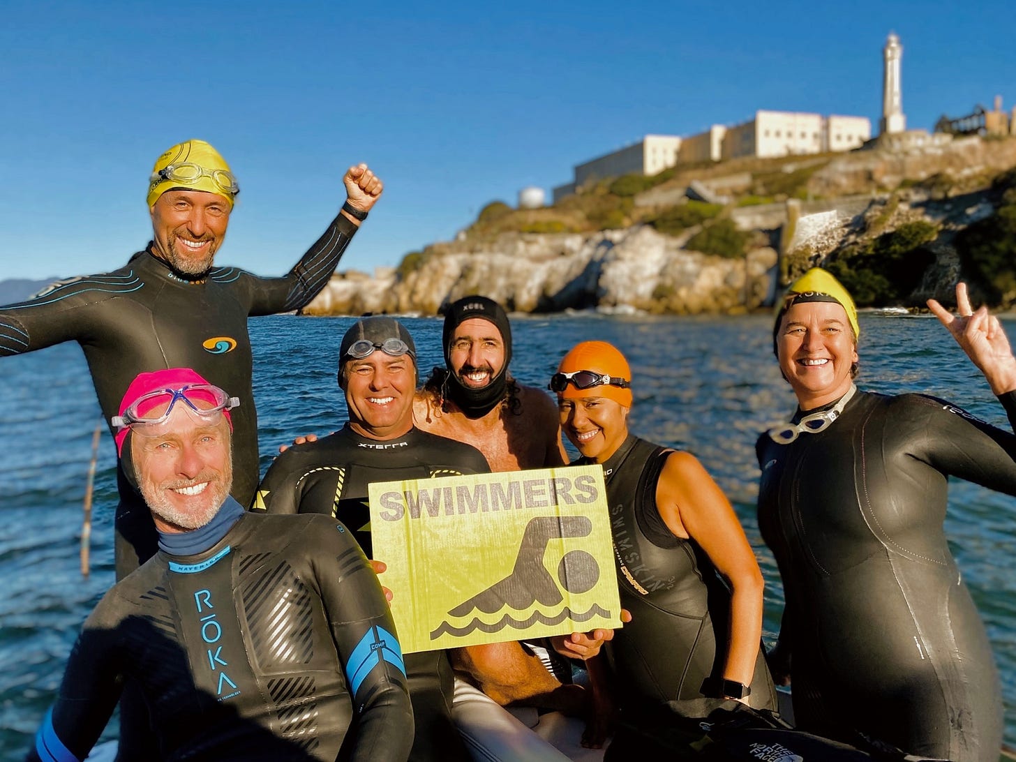 Group of six open water swimmers in wetsuits posing on a boat in front of Alcatraz Island, holding a “Swimmers” sign before a successful cold water swim.