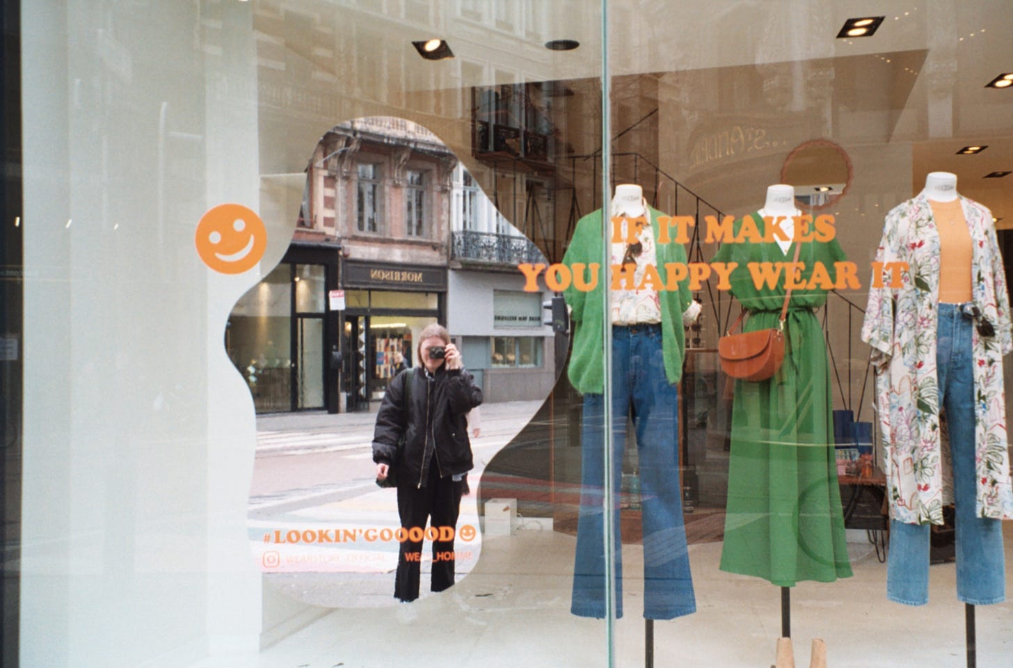 A film snapshot of my reflection in a shop window—dressed in black, surrounded by vibrant colours and smiley faces. The hashtag #lookin’goood adds a playful touch.