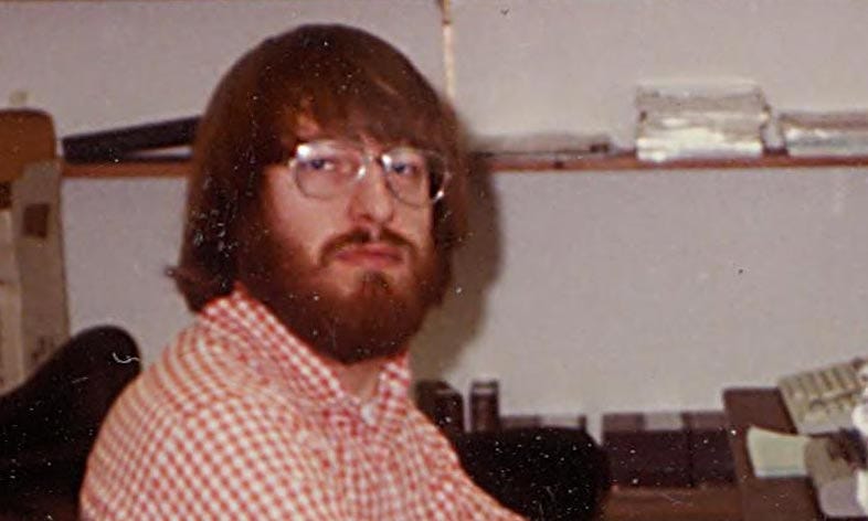 1980s photo of Steve Meretzky sitting at a computer. He wears a checked collared shirt, chunky eyeglasses, and a hairdo and beard that could be called “Wookiee-esque.”