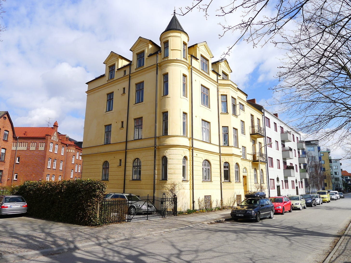 A large buuilding, four stories high,shown at the corner of a street. The building has small towers and was built at the start of the 20th century
