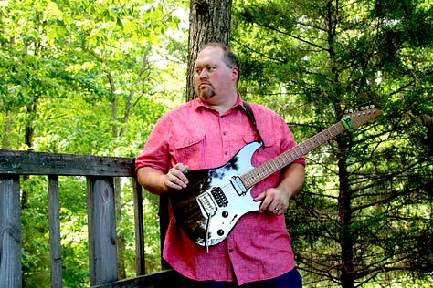 Eric Beaty playing guitar posing with Breedlove and Ibanez guitars wearing a NaNoWriMo winner shirt and posing with Smart Focus book series